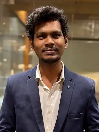 Portrait of a man with dark hair wearing a navy blazer over a white collared shirt, standing indoors with a blurred background.