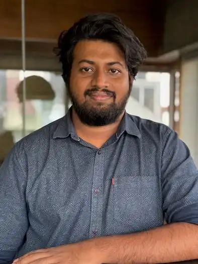 Young man with dark hair and beard, wearing a blue patterned shirt, smiling and sitting indoors with blurred background.
