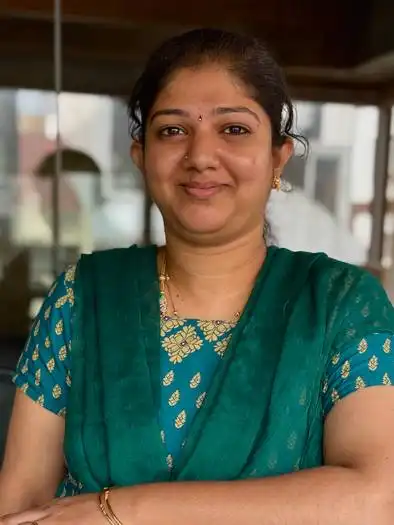 Smiling woman with dark hair tied back wearing a teal traditional outfit with gold patterns, standing indoors with arms crossed.