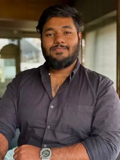 Portrait of a bearded man with short black hair wearing a dark button-up shirt and a wristwatch, standing indoors with arms crossed and a slight smile.