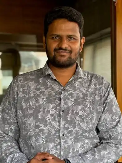 Man with short black hair and beard wearing a gray floral patterned shirt, standing indoors with hands clasped, smiling slightly.