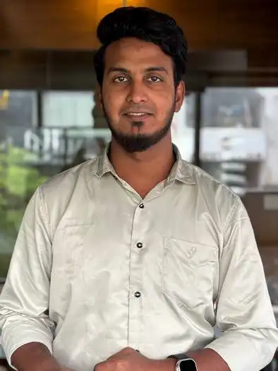 A man with dark hair and a short beard wearing a white button-up shirt and a smartwatch, standing indoors in front of a large window with a blurred outdoor view.