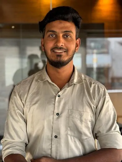 Young man with dark hair and beard wearing a light-colored, buttoned shirt standing indoors with a warm-toned wooden wall and glass panels in the background