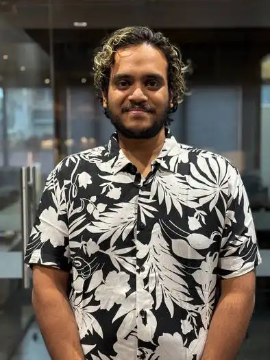 A man with short curly hair wearing a black and white floral patterned shirt standing indoors with glass doors in the background.