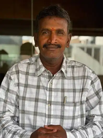 Portrait of a middle-aged man with a mustache wearing a white and gray plaid shirt, standing indoors with blurred background and hands clasped in front.