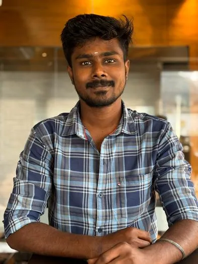 Portrait of a man with dark hair and beard, wearing a blue and white checked shirt, sitting with folded arms indoors against a warm-toned background.