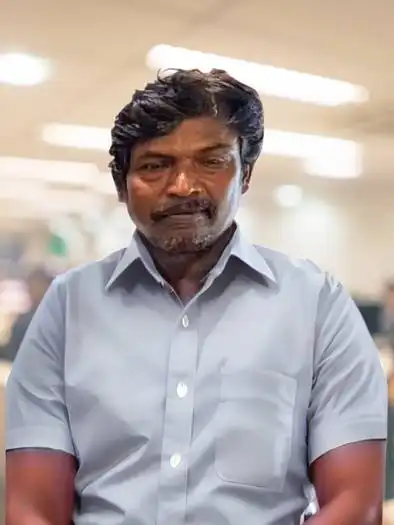 Portrait of a man with dark hair and beard wearing a light blue button-up shirt, standing indoors with blurred background lights.