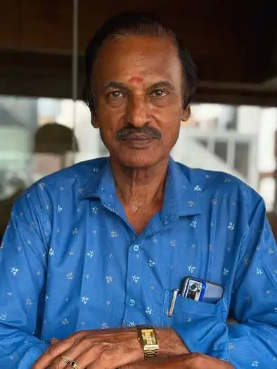 Middle-aged man with a mustache wearing a blue patterned shirt, a watch, and a ring, seated indoors with a smartphone and pen visible in his shirt pocket.