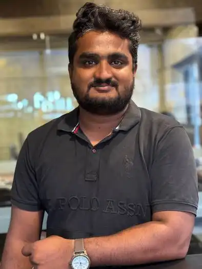 Man with dark hair and beard wearing a dark polo shirt with a watch on his left wrist, standing indoors with arms crossed and a blurred background.