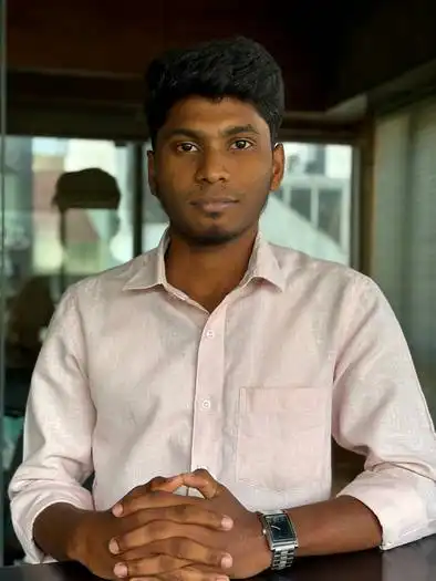 Young man with short black hair wearing a light pink button-up shirt and a wristwatch, sitting at a table with his hands clasped, in an office setting with glass walls and blurred background.