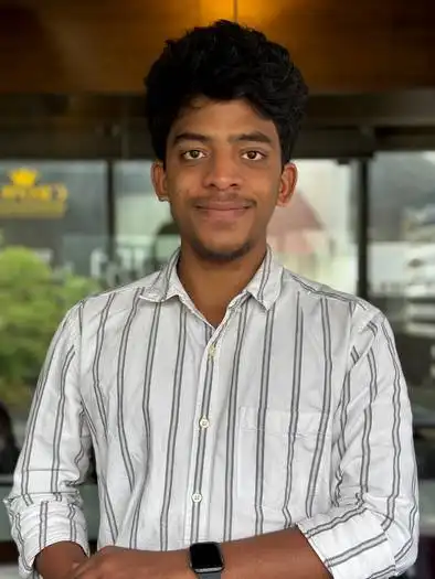 Young man with curly black hair wearing a white button-up shirt with black vertical stripes, standing indoors with blurred windows and greenery in the background, smiling at the camera.