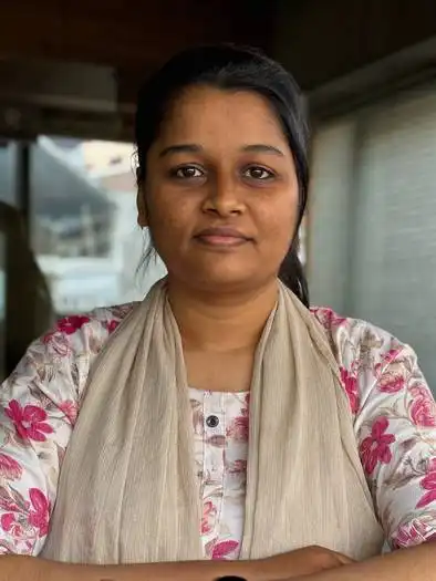 Portrait of a woman with dark hair tied back, wearing a beige scarf over a white floral patterned top, standing indoors with her arms crossed and a neutral expression.
