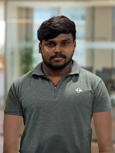 Portrait of a man with short black hair and beard, wearing a dark green polo shirt, standing indoors with a blurred office background.