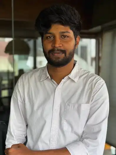 Portrait of a young man with dark hair and beard wearing a white button-up shirt, standing indoors with blurred background.