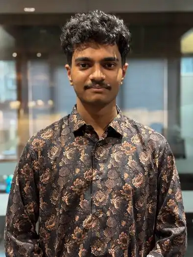 Young man with curly hair and a mustache wearing a dark floral patterned button-up shirt standing indoors with blurred background