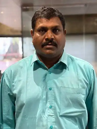 Portrait of a middle-aged man with short black hair and a mustache, wearing a light teal button-up shirt, standing indoors with a blurred window and curtain in the background.