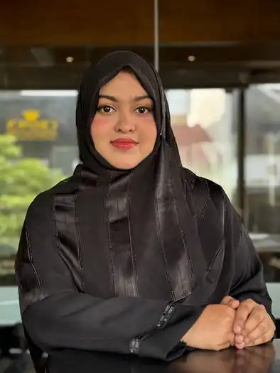 Portrait of a young woman wearing a black hijab and black clothing, sitting indoors with her hands folded on a table, smiling gently at the camera.