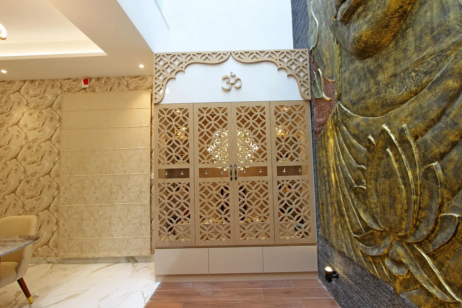 A divine prayer space showcases carved lattice doors, marble flooring, golden Buddha wall art, soft recessed lighting, and an illuminated deity niche radiating spiritual serenity.
