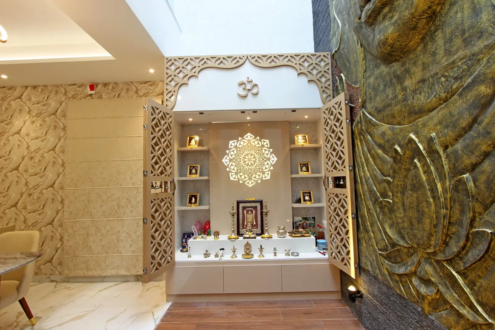 This prayer room features intricately carved lattice doors with an Om symbol, marble and wooden flooring, textured beige walls, and a grand golden Buddha mural radiating spiritual grace.