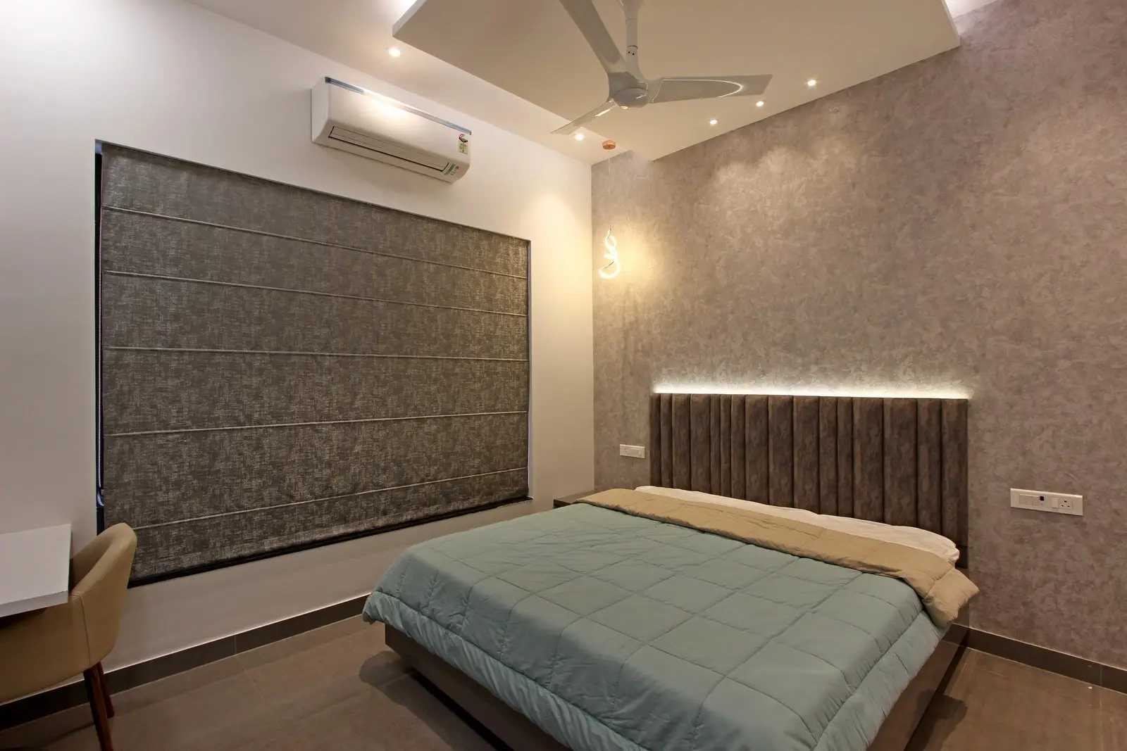 The deep grey velvet headboard with soft LED lighting creates a warm focal point, while Roman-shaded windows and recessed ceiling lights enhance comfort and modern light control.


