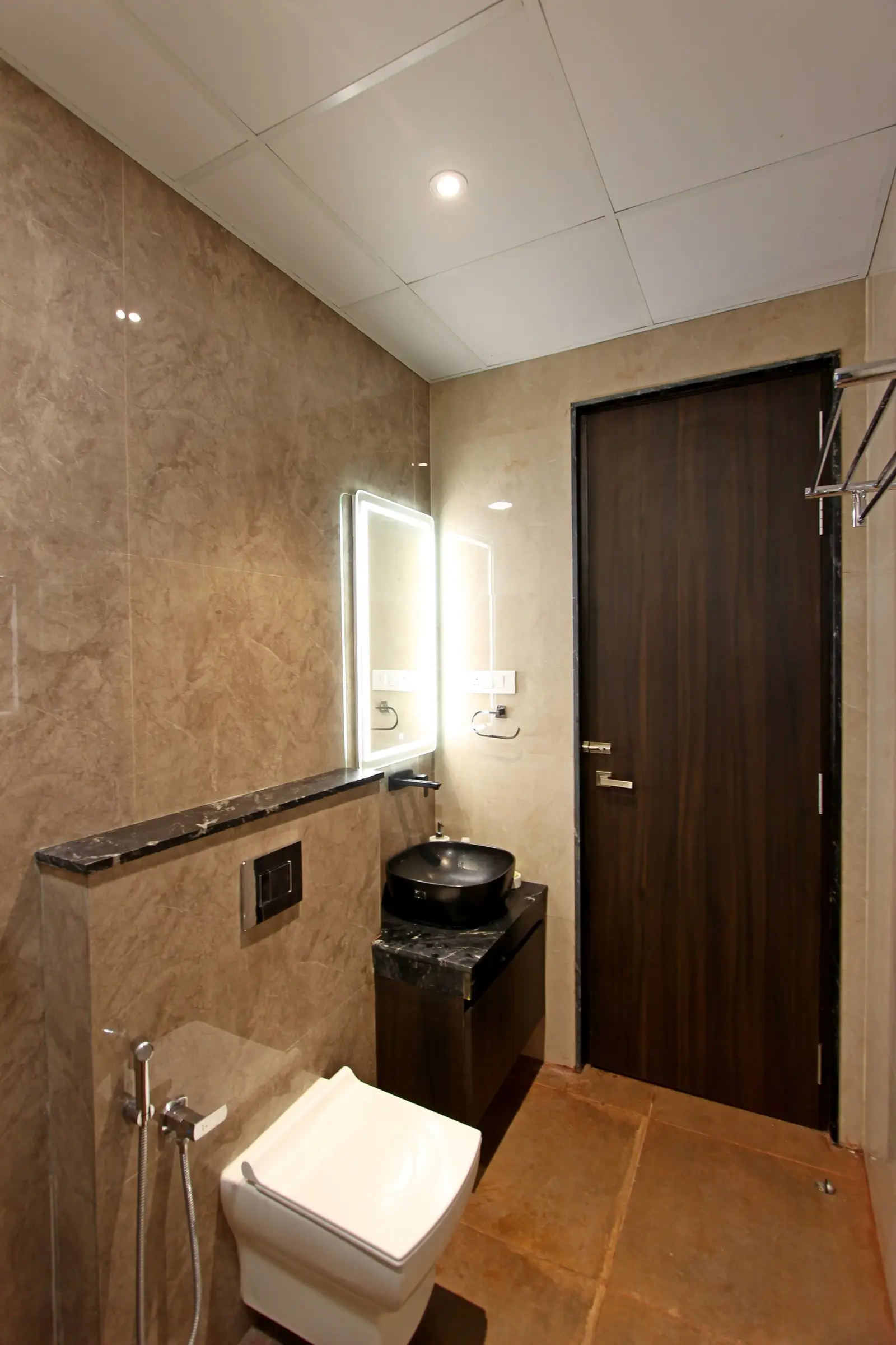 A elegant bathroom features beige marble walls, a wall-mounted toilet, a black granite vanity with a matte vessel sink, and an illuminated mirror enhancing sophistication.

