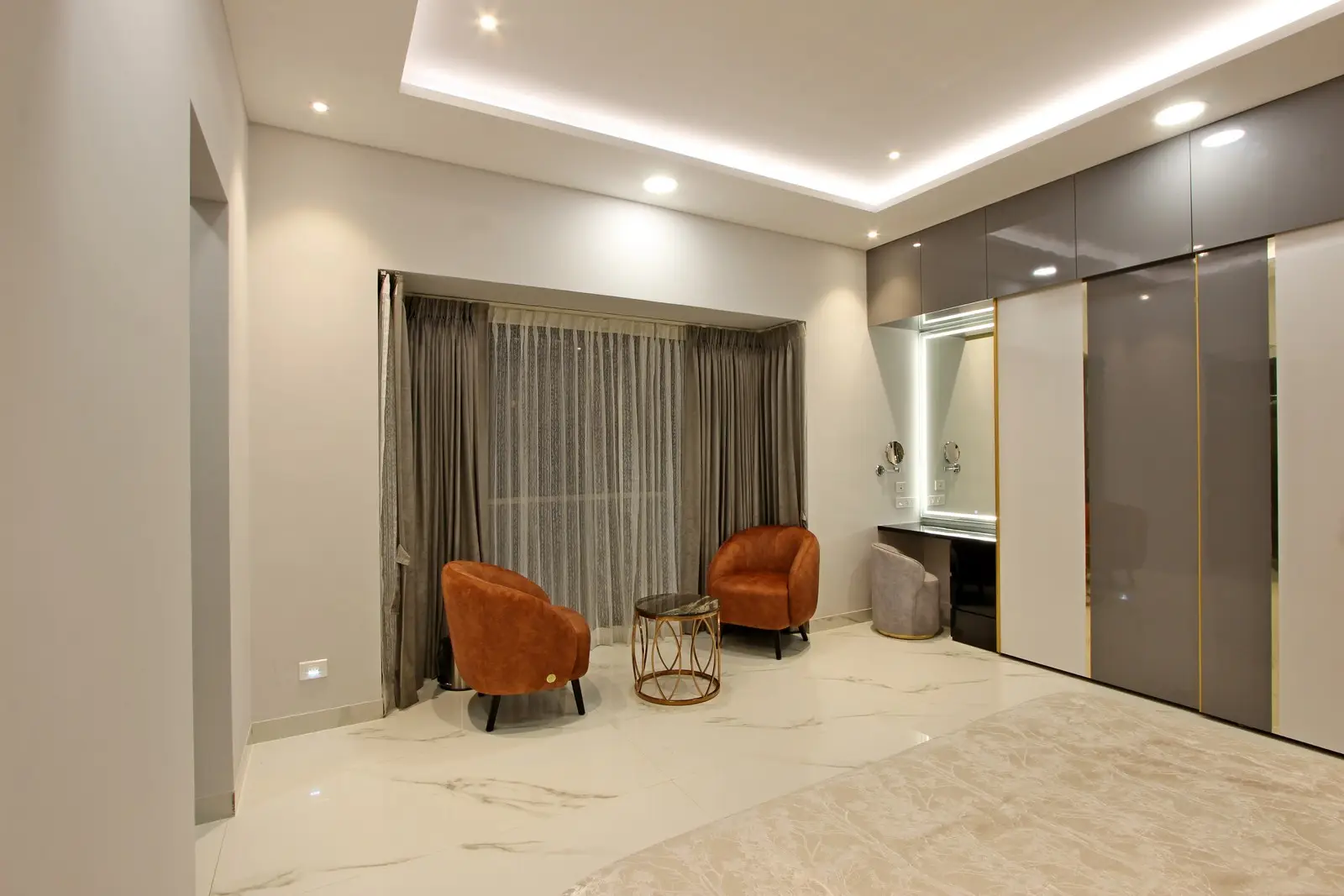 Near the large window, sheer curtains frame a cozy seating nook featuring a warm brown accent chair and a metallic round table.
