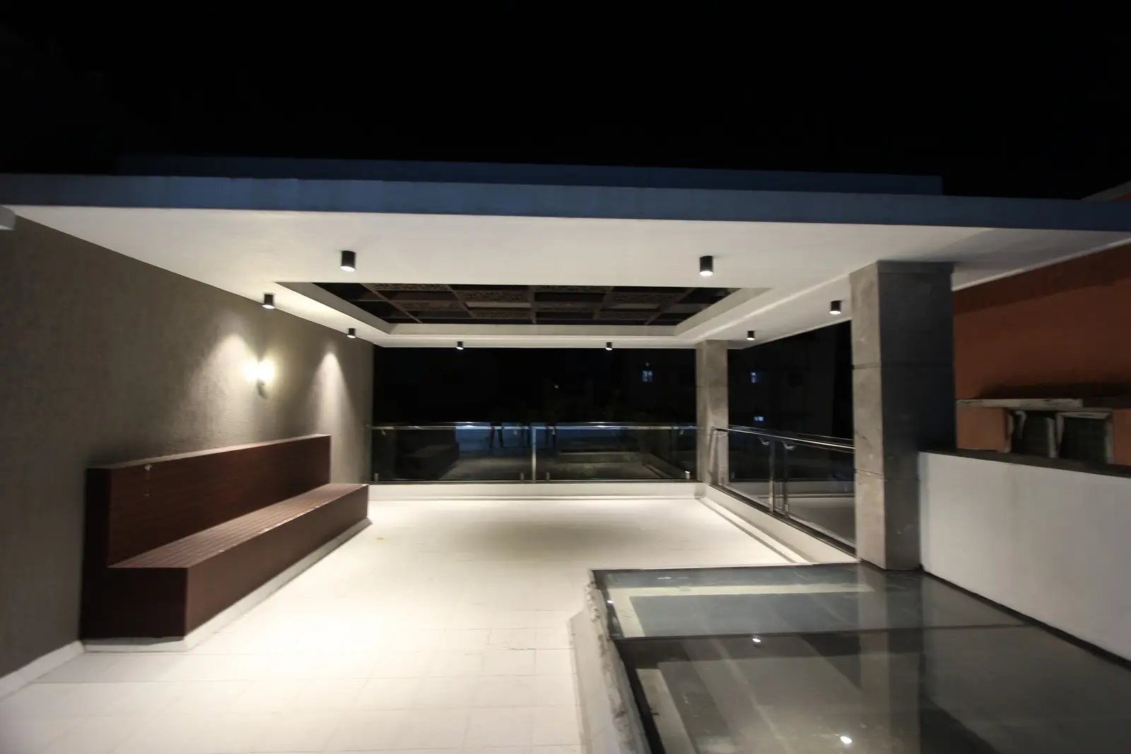 This modern terrace combines white tiles, recessed lighting, glass railings, and a wooden bench to create an elegant, open, contemporary space for relaxing gatherings.