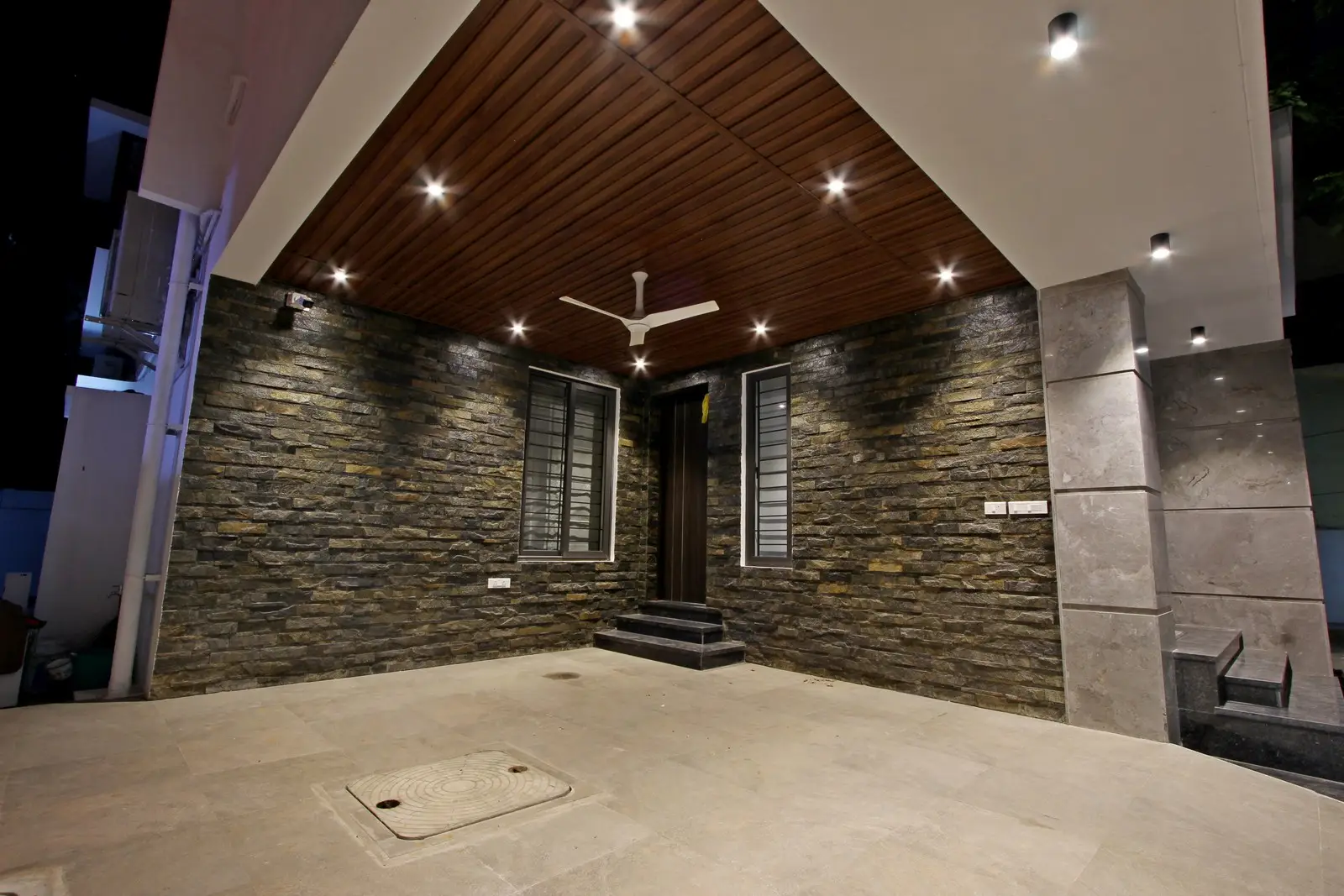 This exterior features stone-clad walls, a wooden ceiling with recessed lighting, and sleek grey flooring, creating a warm, elegant, and contemporary entrance design.