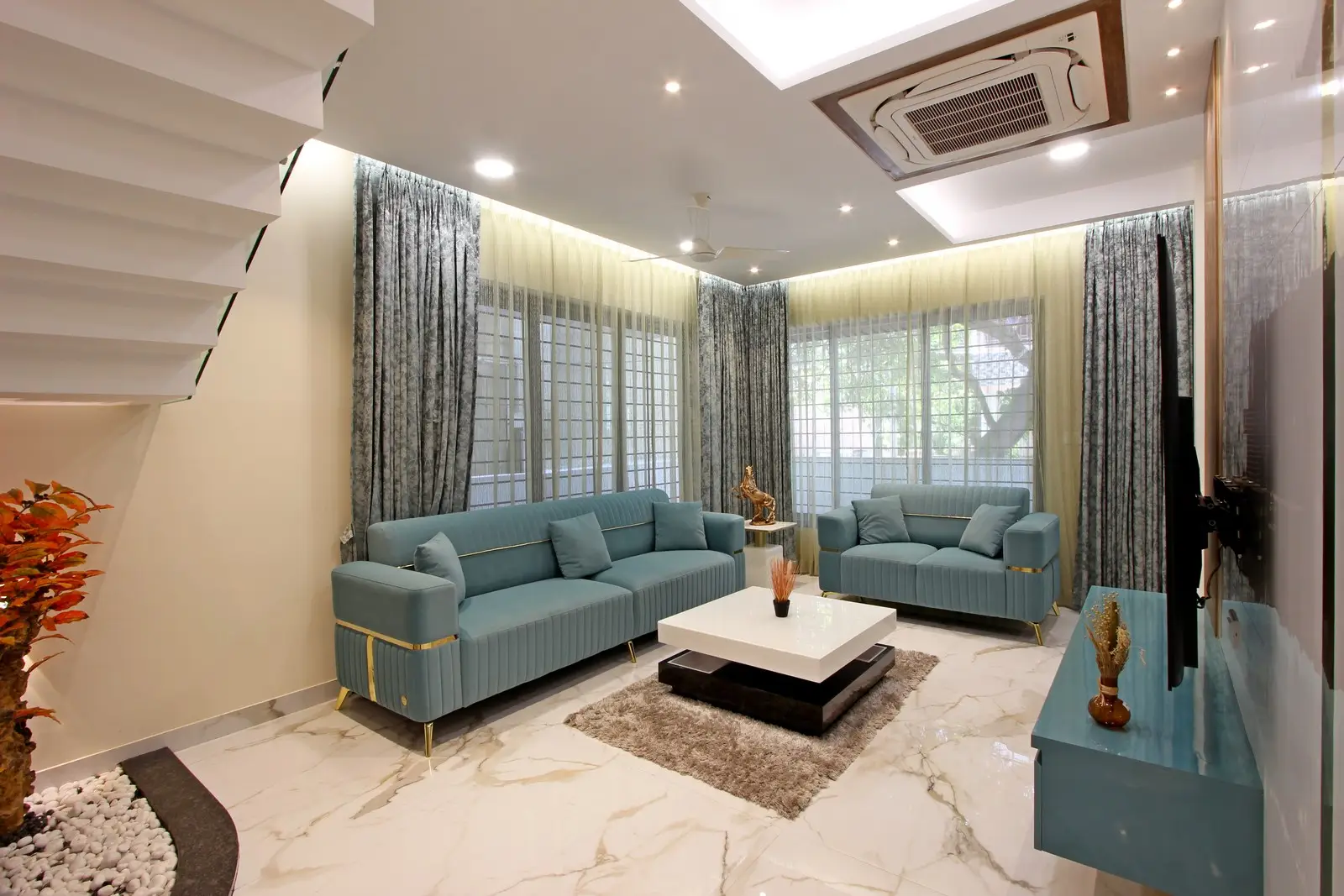 Modern living room with two blue sofas, a white and black square coffee table on a brown rug, sheer and dark curtains covering large windows, marble floor, a mounted TV on the right wall above a blue floating cabinet, and a staircase on the left side with plants beneath it.