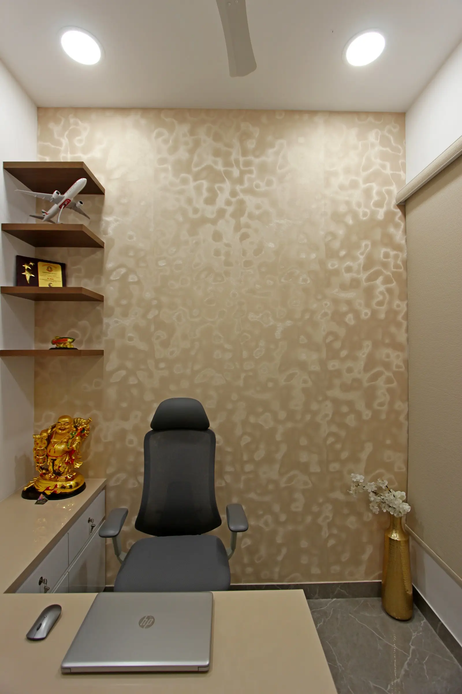 A modern workspace featuring a textured beige wall, sleek grey chair, marble flooring, and wooden shelves with golden décor, creating a refined professional atmosphere
