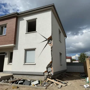 How to Stop Cracks in Your Home: Professional Guidance from Architect Dameem Ansari 12 Two-story house with a large vertical crack running from near the roof to the ground on the white side, exposing broken bricks and rubble at the base, with scattered debris around the house exterior under a partly cloudy sky.