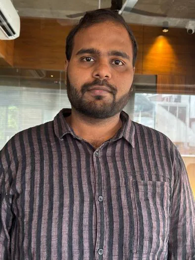 Young man with a beard wearing a vertically striped button-up shirt standing indoors with a wooden wall and glass window in the background.