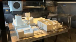 White architectural model or prototype being 3D printed inside a 3D printer with the printing nozzle visible above the model.