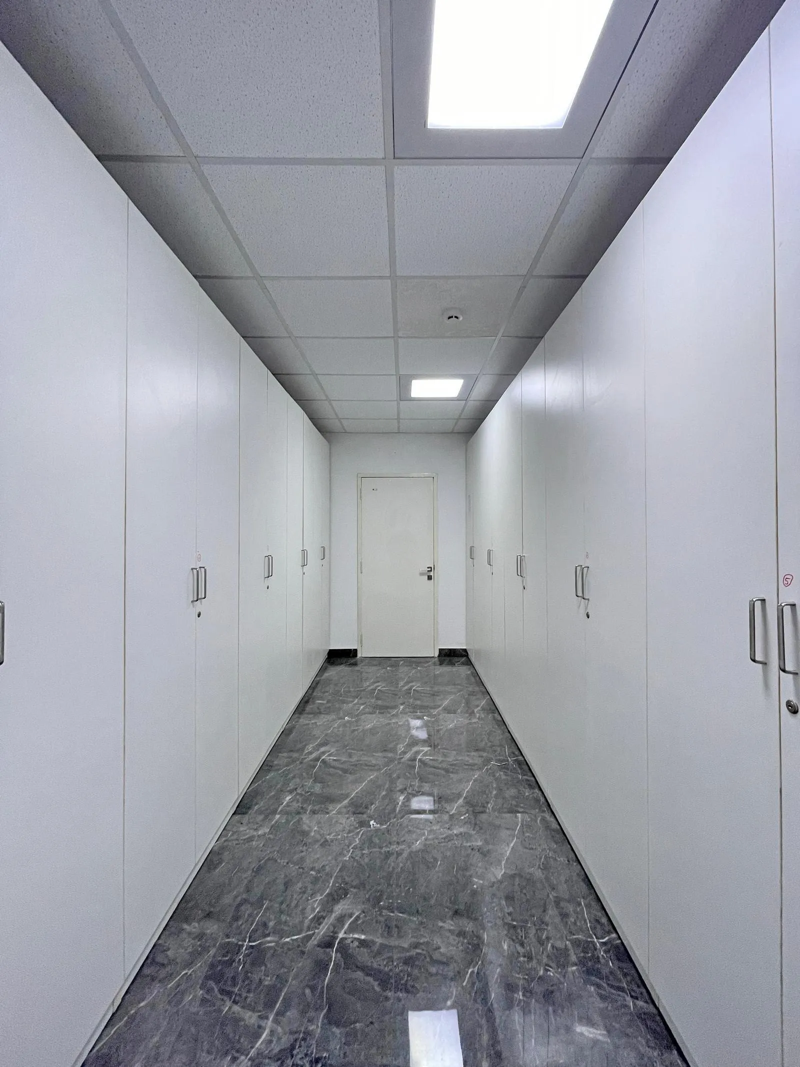 The space features full-height modular storage, high-gloss marble flooring, and a suspended acoustic ceiling with recessed lighting for a sleek, functional design. A linear layout and clean white finishes enhance circulation, efficiency, and professional aesthetics.