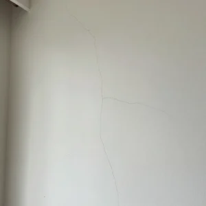 How to Stop Cracks in Your Home: Professional Guidance from Architect Dameem Ansari 9 White wall with multiple thin, irregular vertical and horizontal cracks running through it near a corner with a ceiling and curtain rod partially visible