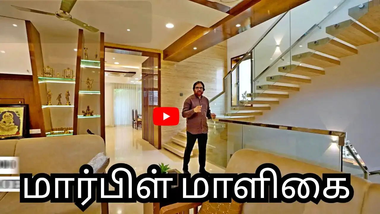 Man standing inside a modern home interior with floating wooden stairs and glass railing, visible living room furniture, and decorative shelves with brass figurines, with Tamil text overlay at the bottom.