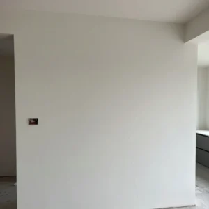 How to Stop Cracks in Your Home: Professional Guidance from Architect Dameem Ansari 8 Plain white wall with an empty electrical box opening on the left side and a faint vertical crack running down the right side, adjacent to a corner opening revealing part of a room with a window and a cabinet.