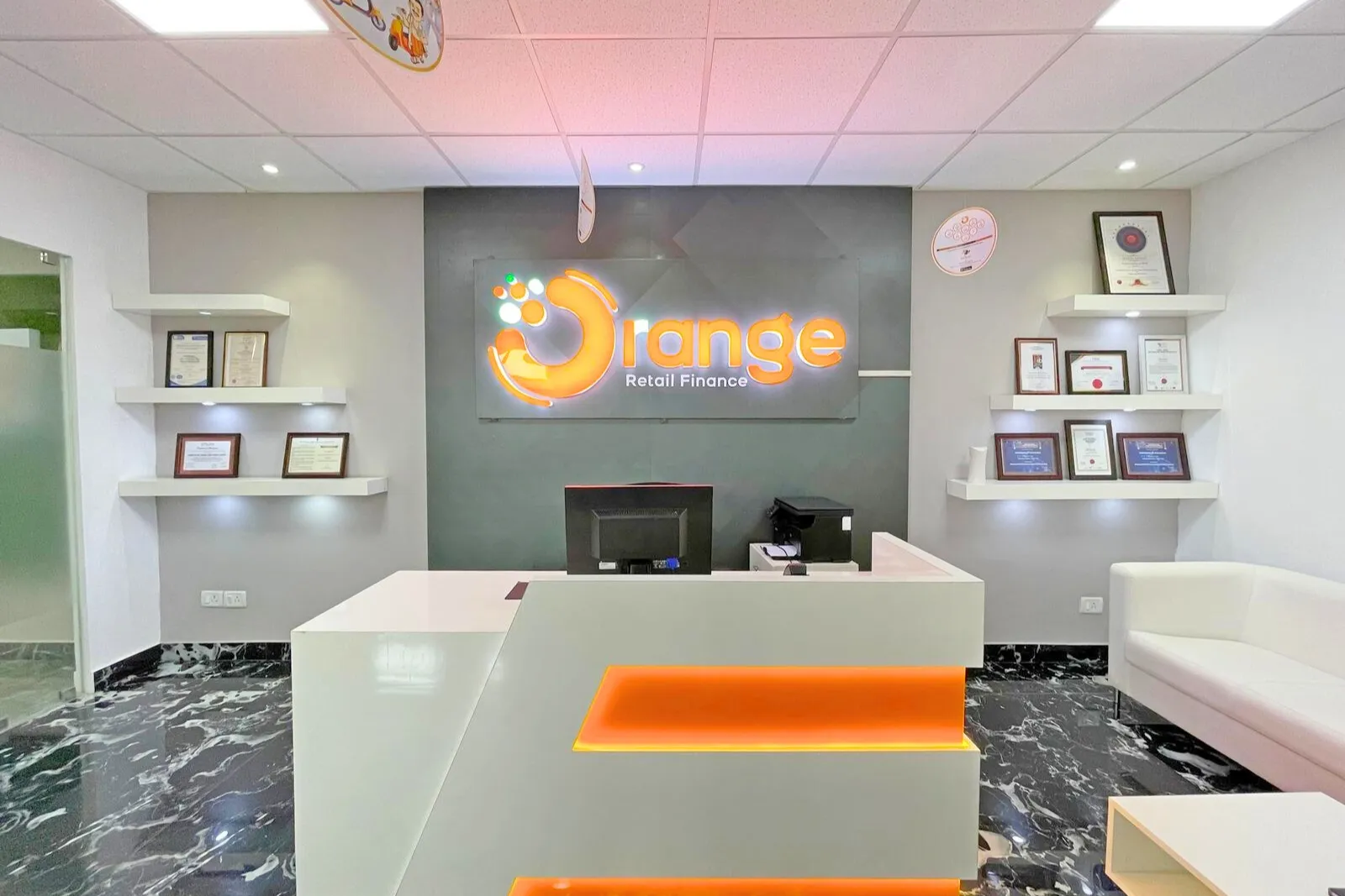 Modern office reception area with a white desk featuring orange LED lighting, a black marble floor, white walls with floating shelves displaying certificates and awards, a white sofa, and an illuminated Orange Retail Finance logo on a dark gray wall behind the desk.