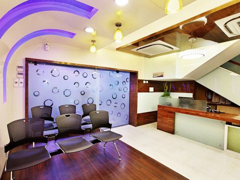 Modern waiting area with six black chairs arranged on a wooden floor, frosted glass wall with circular patterns, illuminated ceiling lights, and a wooden reception desk under a staircase.