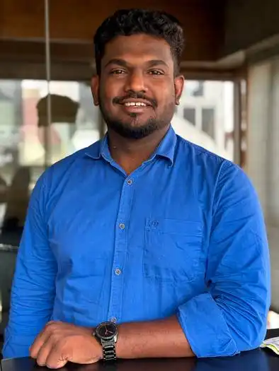 Smiling man with short curly hair and beard wearing a blue button-up shirt and a black wristwatch, standing indoors with his arm resting on a surface.