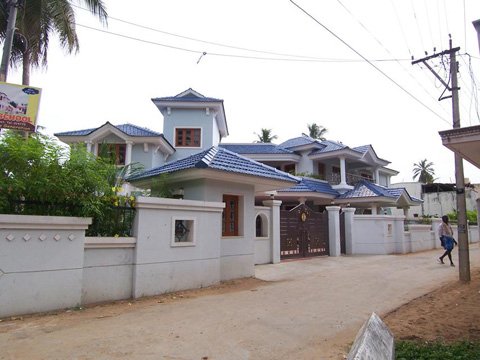 Large white house with blue tiled roof behind a white boundary wall along a dirt road with a person walking on the right side