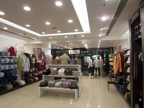 Interior of a well-lit clothing store displaying neatly folded shirts on shelves and racks, with three mannequins dressed in casual wear near the back under signs reading 'Genesis' and 'Basics'