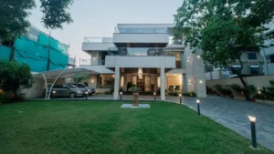 Modern multi-story house with large entrance, glass balconies, and landscaped front yard with green grass and illuminated pathway lights, cars parked under a white canopy to the left, and trees framing the scene.