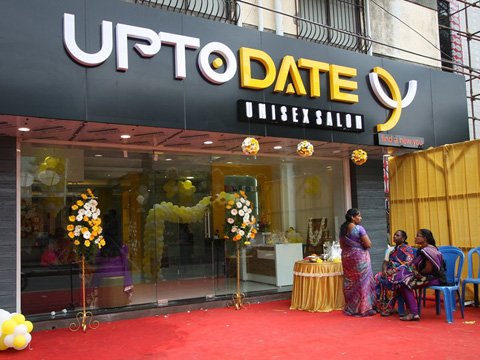 Exterior of a unisex salon named 'UP TO DATE' with a black and yellow signboard, decorated with flower stands and balloons, and three women conversing near the entrance on a red carpeted area.