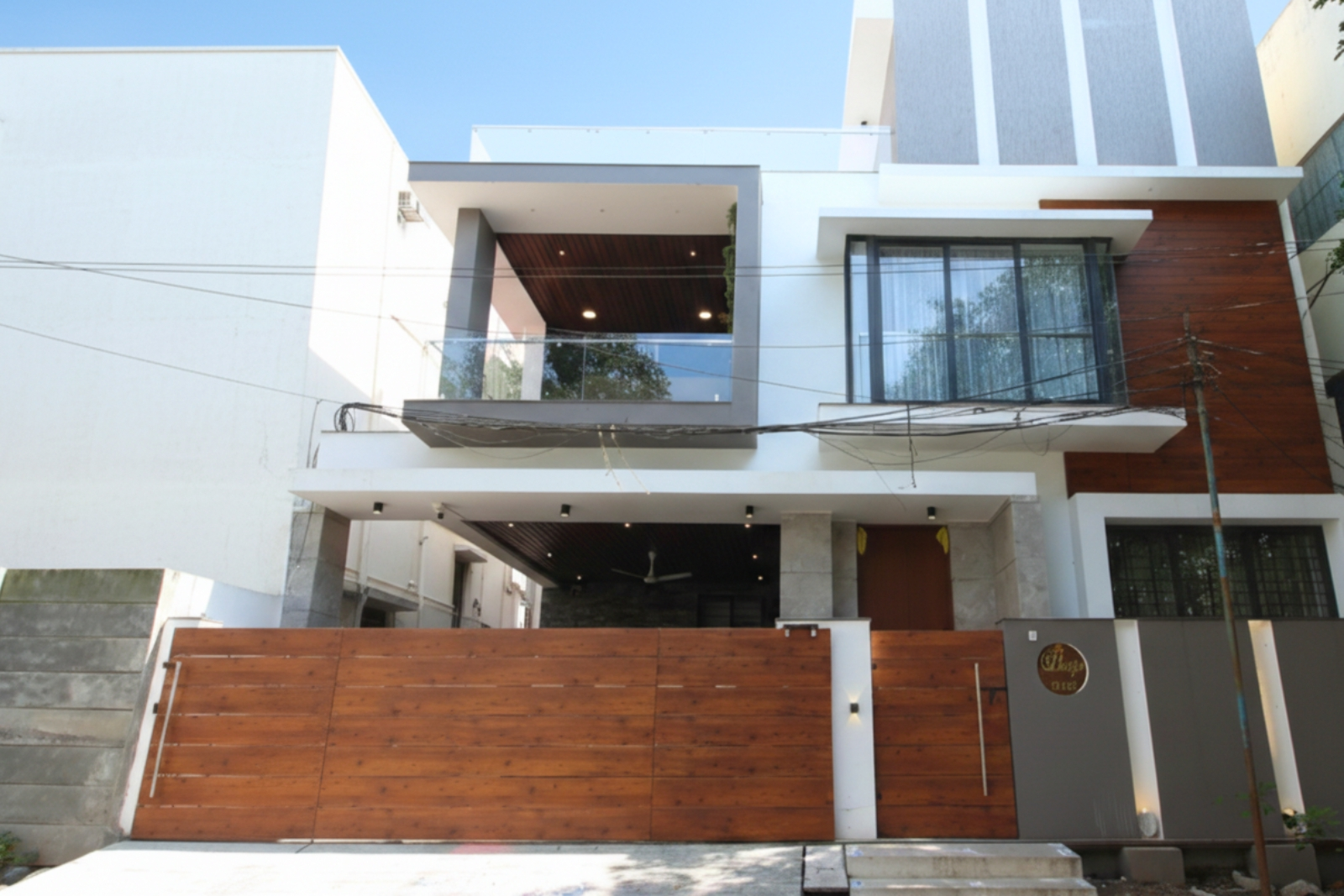 Modern two-story house with a white and gray exterior, large wooden gate, and a balcony with glass railing under a blue sky.
