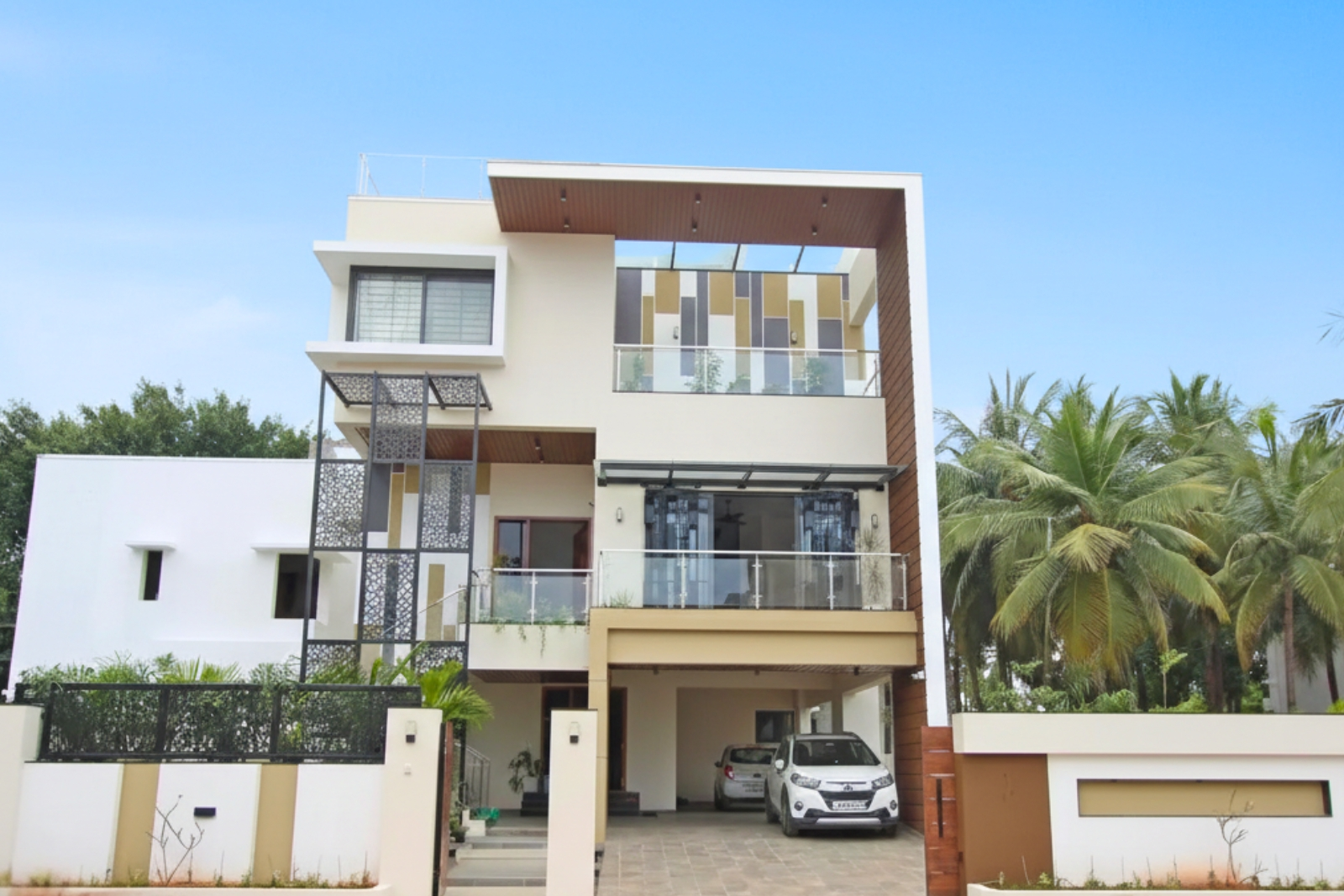 Modern two-story house with a beige and white exterior, large windows, metal decorative panels, glass balcony railings, a covered carport with two cars, and palm trees in the background under a clear blue sky.