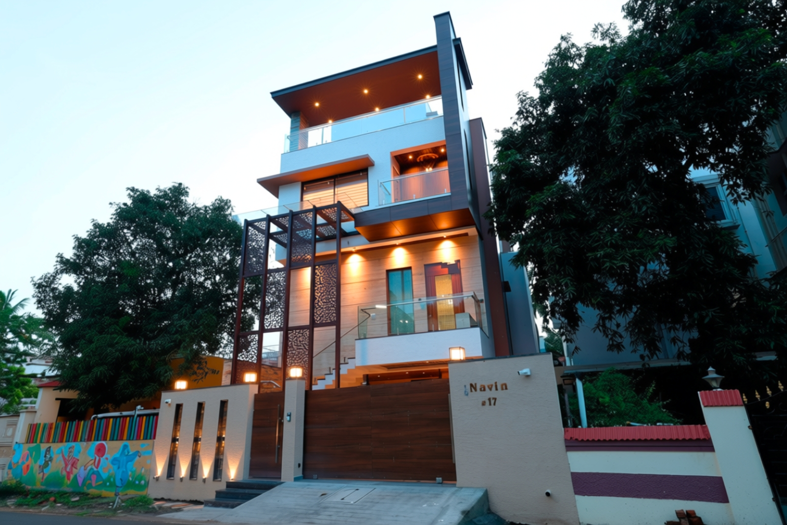 Modern three-story house with warm exterior lights, wooden accents, and glass balcony railings, surrounded by large leafy trees and a colorful mural on the adjacent boundary wall.