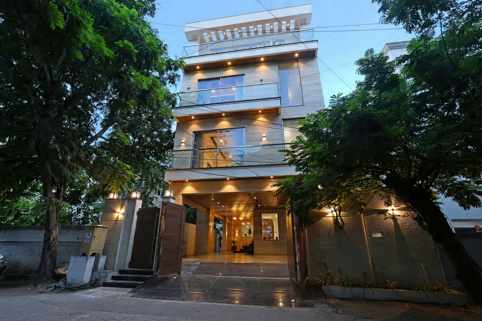 Modern multi-story building with large glass windows and balconies, illuminated exterior lights, open wooden gate, and surrounding trees at dusk.