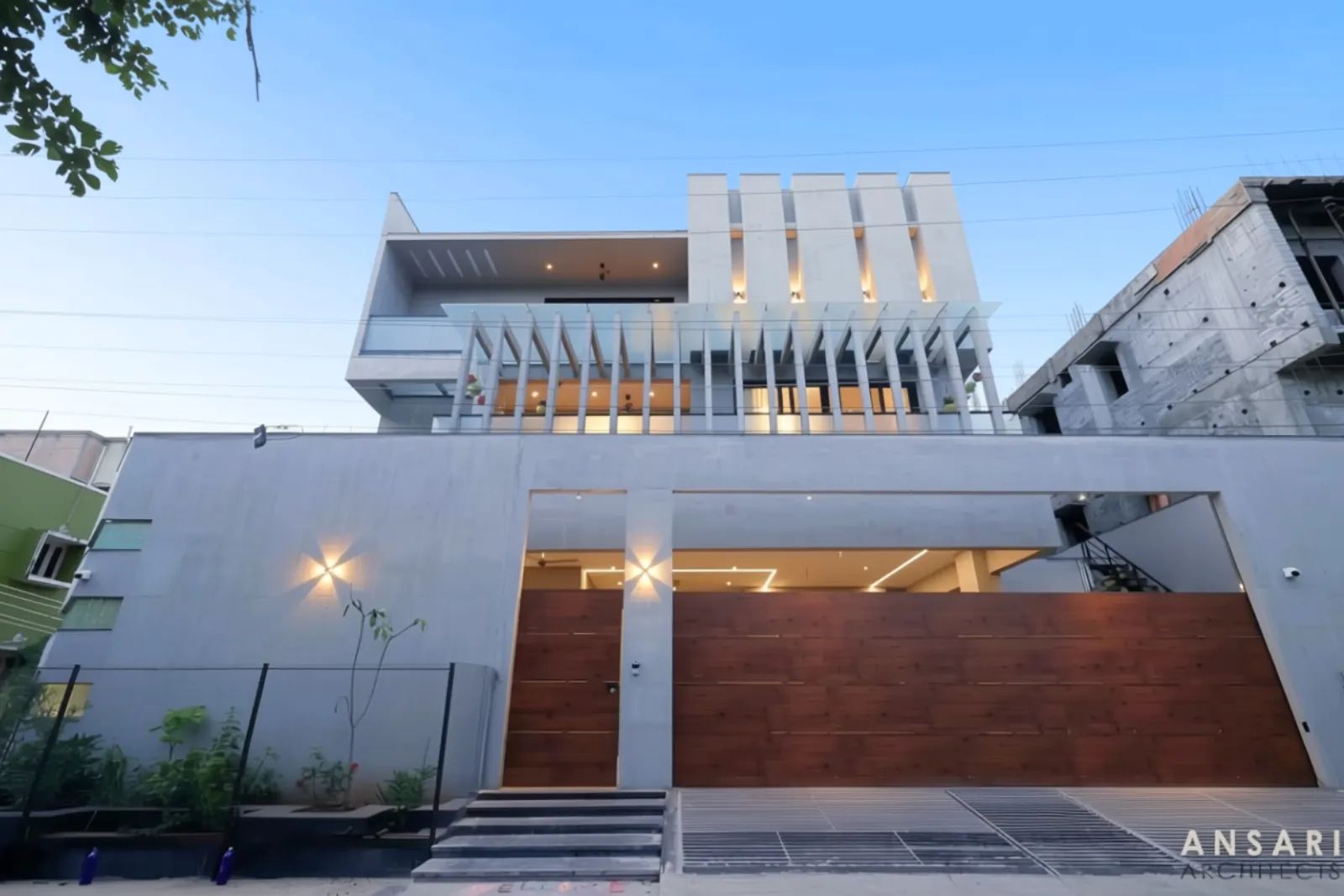 Modern multi-story gray concrete house with wooden gate, decorative vertical panels, warm exterior lights, and a clear blue sky background.