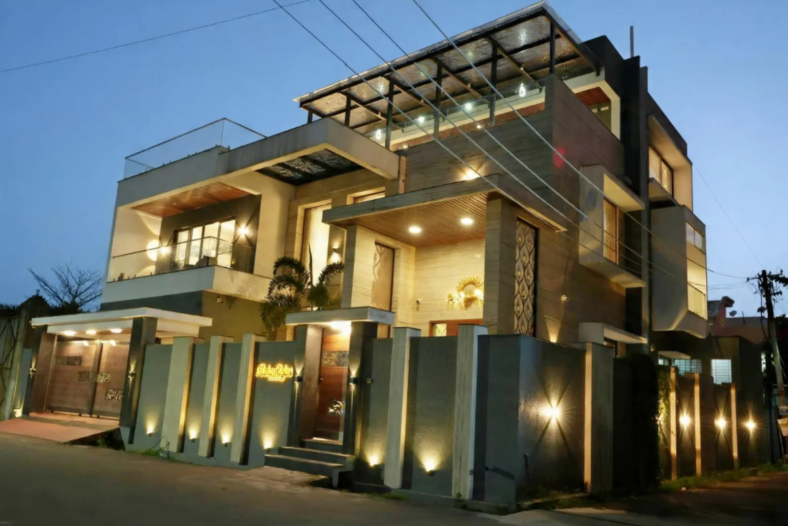 Modern two-story house with large balconies and warm exterior lighting at dusk, featuring a tall fence and trees near the entrance.